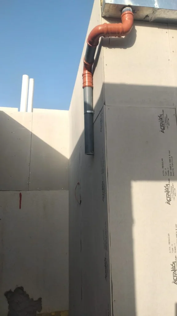 جدار خارجي مكسو بألواح أسمنتية ليفية تظهر عليها بطاقات المنتج المطبوعة، مع ماسورتَي تصريف رأسيتين برتقالية ورمادية وحافة السقف أعلاه