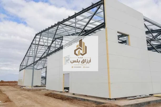 هيكل معدني بجمالونات ظاهرة أثناء كسوة جدرانه بألواح معزولة بيضاء مع فتحات نوافذ محدّدة الإطار