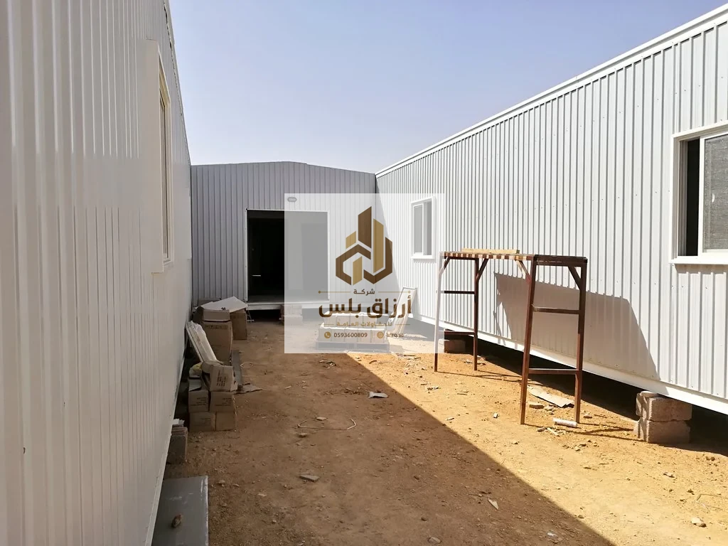 ممر ضيّق بين مبنيين مكسوّين بألواح معزولة مضلّعة مع باب معدني ومواد موضوعة على أرض رملية