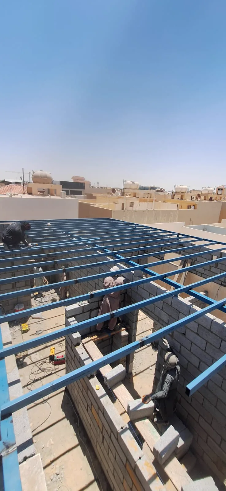 عامل يثبّت مدادات معدنية زرقاء فوق جدار بلوك تمهيدًا لتركيب ألواح السقف