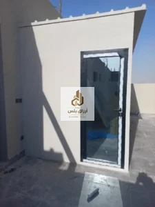 غرفة ساندوتش بانل على سطح، بابها من الألمنيوم والزجاج وتظهر حافة السقف المائلة أعلى الجدار