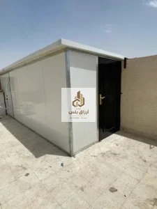 غرفة ساندوتش بانل بيضاء منفّذة داخل فناء، بابها المعدني مفتوح ويظهر جدار الفناء خلفها