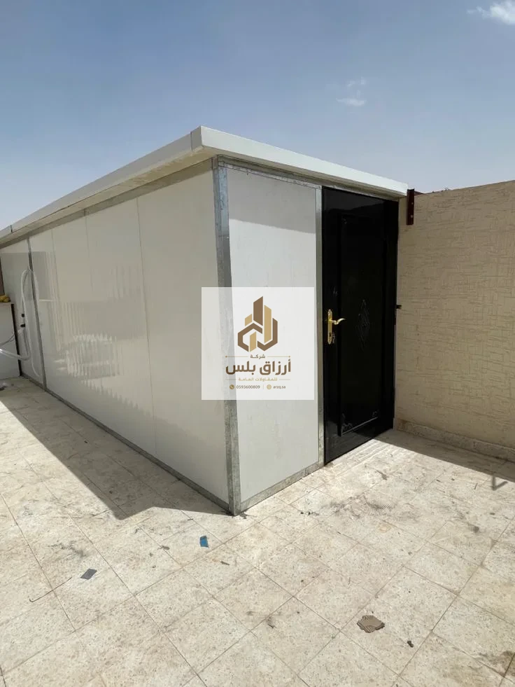 غرفة ساندوتش بانل بيضاء منفّذة داخل فناء، بابها المعدني مفتوح ويظهر جدار الفناء خلفها