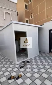 غرفة ساندوتش بانل بيضاء قائمة في فناء مبلّط ببلاط رمادي وأبيض، وفتحة بابها واسعة