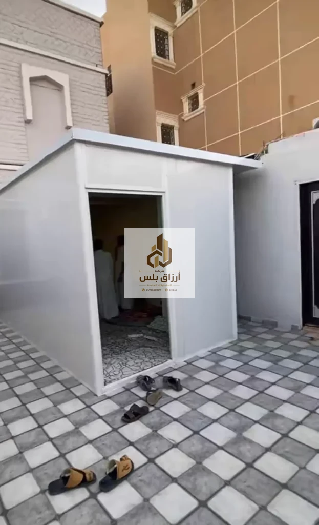 غرفة ساندوتش بانل بيضاء قائمة في فناء مبلّط ببلاط رمادي وأبيض، وفتحة بابها واسعة