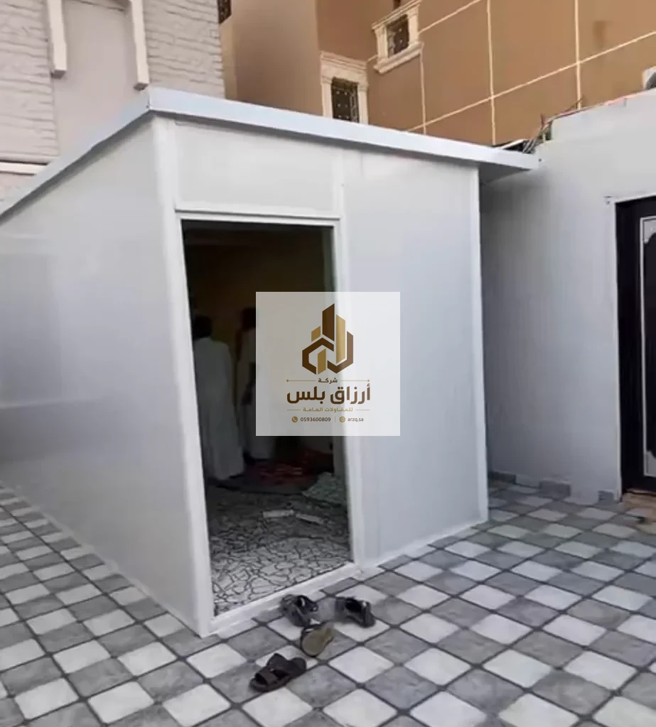 غرفة ساندوتش بانل بيضاء قائمة في فناء مبلّط ببلاط رمادي وأبيض، وفتحة بابها واسعة