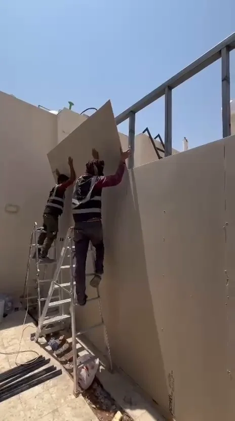 عاملان على سلّم يرفعان لوح ساندوتش بانل كبيرًا لتثبيته ضمن جدار الغرفة